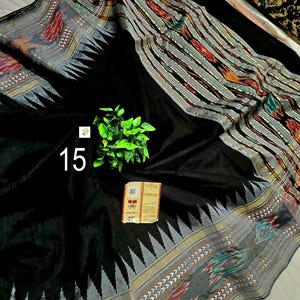 May include: A black saree featuring a wide border with intricate ikat patterns in red, green, and grey, accented by a sharp triangular motif. The fabric displays a solid black center, a small green plant sprig, and a rectangular tag with the number 15.