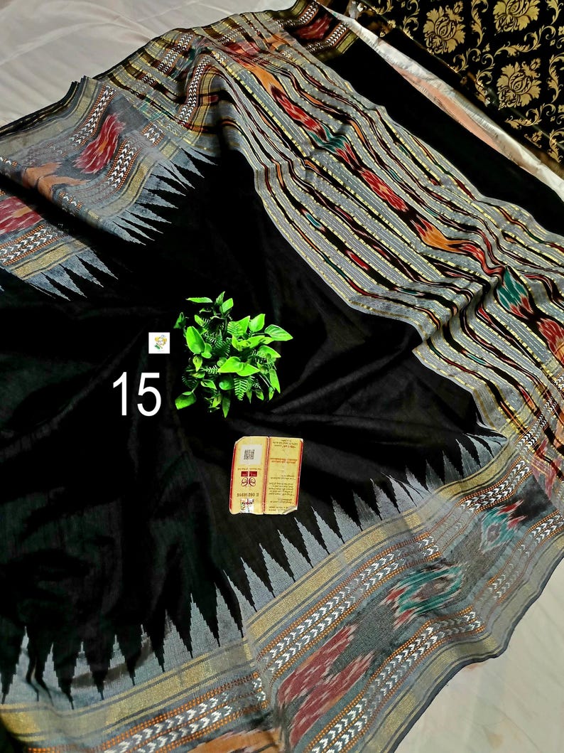 May include: A black saree featuring a wide border with intricate ikat patterns in red, green, and grey, accented by a sharp triangular motif. The fabric displays a solid black center, a small green plant sprig, and a rectangular tag with the number 15.