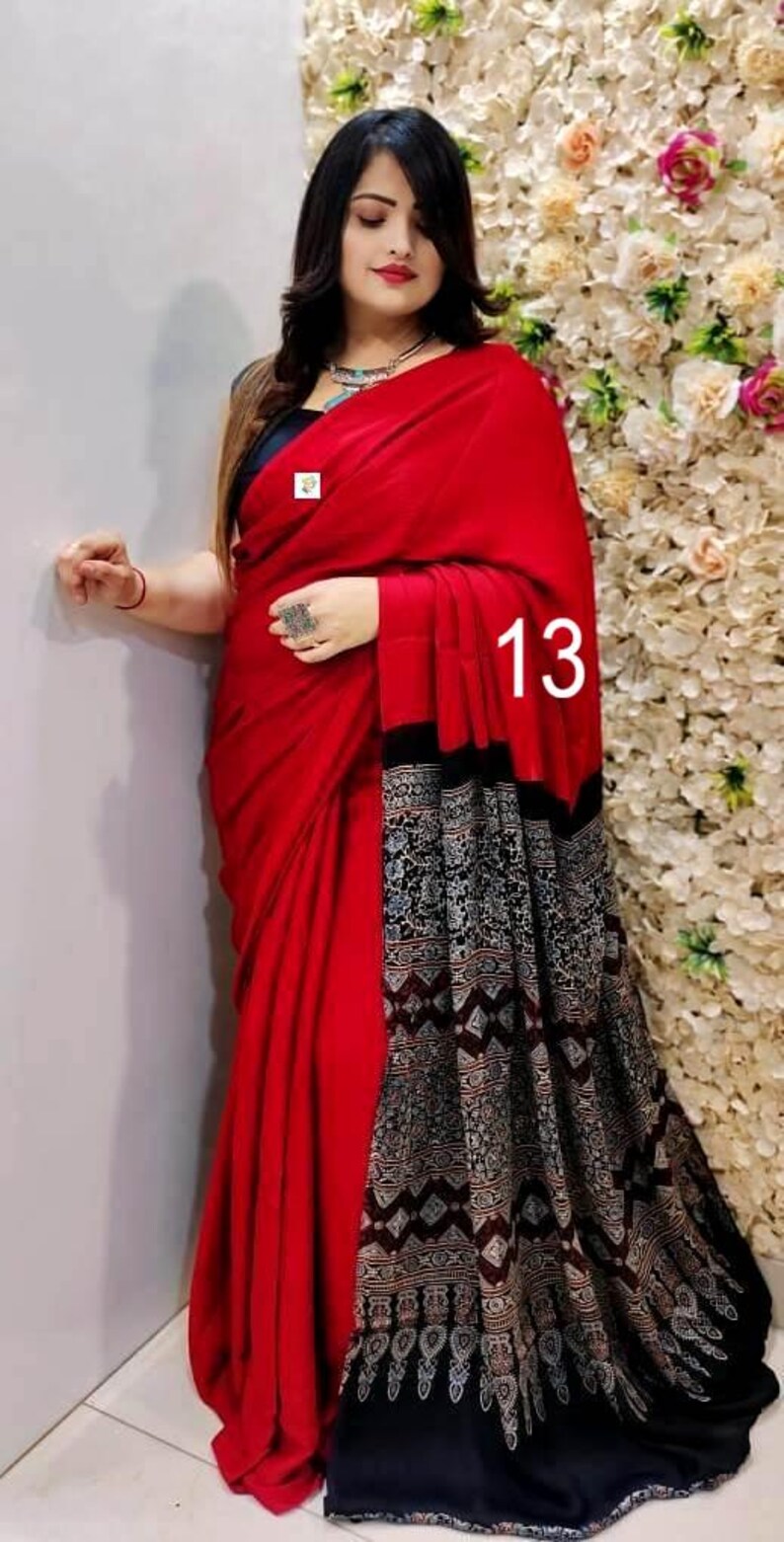 Puede incluir: Un sari de color rojo vibrante con un cuerpo rojo s&oacute;lido y un pallu negro en contraste adornado con intrincados patrones geom&eacute;tricos y florales en blanco y gris. La tela se drapea elegantemente, mostrando una combinaci&oacute;n audaz y el n&uacute;mero 13.