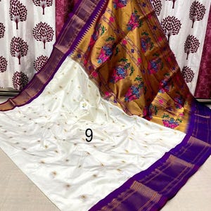 Op de afbeelding: Een witte sari met kleine goud geborduurde bloemmotieven over het hele lichaam, een brede paarse rand met gouden geometrische patronen en een grote gouden pallu versierd met kleurrijke pauwen- en bloemmotieven, gemarkeerd met het cijfer 9.
