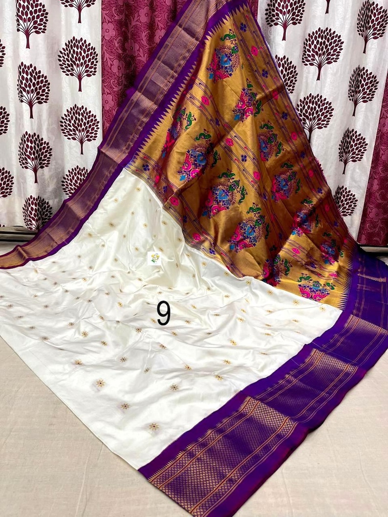 Op de afbeelding: Een witte sari met kleine goud geborduurde bloemmotieven over het hele lichaam, een brede paarse rand met gouden geometrische patronen en een grote gouden pallu versierd met kleurrijke pauwen- en bloemmotieven, gemarkeerd met het cijfer 9.