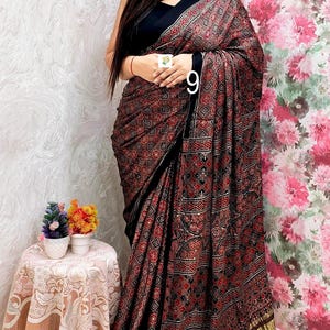 May include: A black saree featuring a dense, repeating geometric and floral pattern in shades of red, white, and black. The garment includes a wide, metallic gold border at the hem and a solid black blouse with a high neckline and a choker necklace.