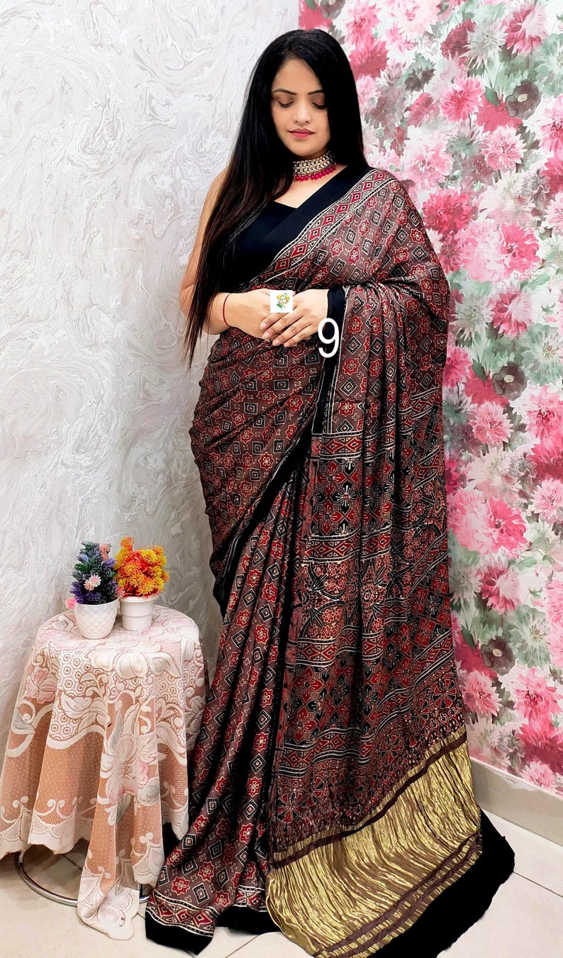 May include: A black saree featuring a dense, repeating geometric and floral pattern in shades of red, white, and black. The garment includes a wide, metallic gold border at the hem and a solid black blouse with a high neckline and a choker necklace.