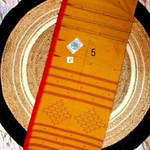 May include: A mustard yellow textile with a vibrant red border and fringe. The fabric features horizontal red stripes, small red dots, and two large diamond-shaped patterns made of red dots. A small white tag displays the number 5 and a logo.