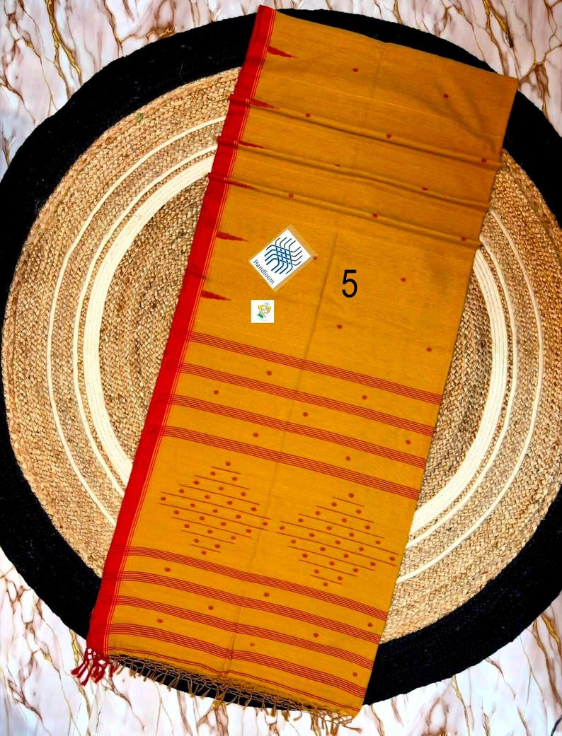 May include: A mustard yellow textile with a vibrant red border and fringe. The fabric features horizontal red stripes, small red dots, and two large diamond-shaped patterns made of red dots. A small white tag displays the number 5 and a logo.