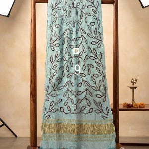May include: A light blue scarf featuring a dark brown embroidered leaf pattern and a metallic gold border at the bottom. The fabric has a crinkled texture and is draped over a wooden stand. The number 9 is printed in white on the center.