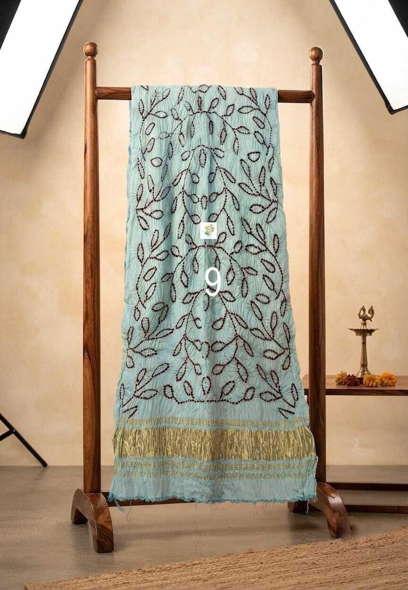 May include: A light blue scarf featuring a dark brown embroidered leaf pattern and a metallic gold border at the bottom. The fabric has a crinkled texture and is draped over a wooden stand. The number 9 is printed in white on the center.
