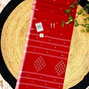 May include: A vibrant red textile featuring white horizontal stripes and small white polka dots. The fabric displays two large diamond-shaped patterns composed of white dots. A small square tag with the text 11 and Handloom is visible.