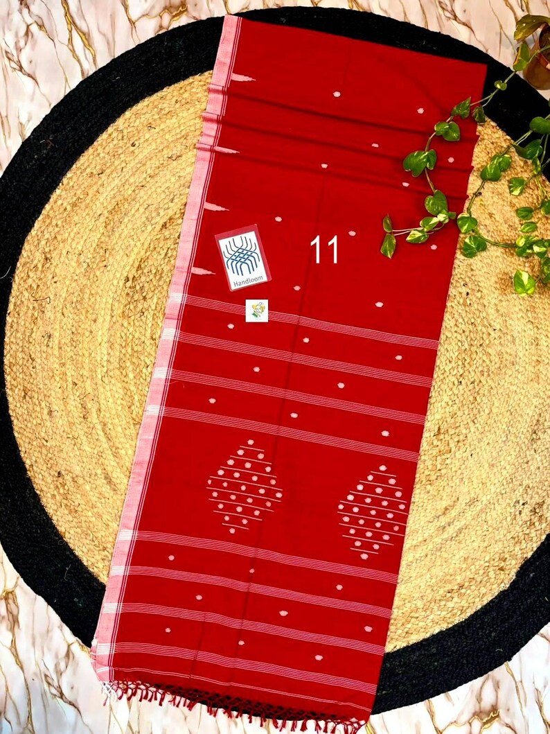May include: A vibrant red textile featuring white horizontal stripes and small white polka dots. The fabric displays two large diamond-shaped patterns composed of white dots. A small square tag with the text 11 and Handloom is visible.