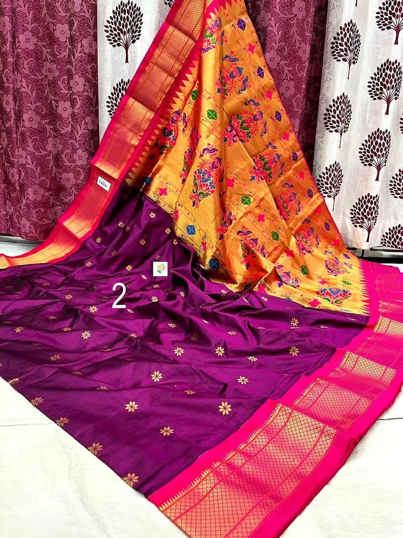 Op de afbeelding: Een dieppaarse sari met kleine gouden bloemmotieven over het hoofdgedeelte, aangevuld met een levendige gouden pallu versierd met kleurrijke pauwen- en bloemmotieven, afgewerkt met een felroze rand en gedetailleerde gouden zari-accenten.