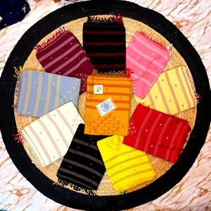 May include: A collection of ten rectangular cotton scarves arranged in a circular pattern. Each scarf features a distinct solid color including maroon, black, pink, light blue, mustard yellow, cream, red, and bright yellow, with thin stripes and small dots.