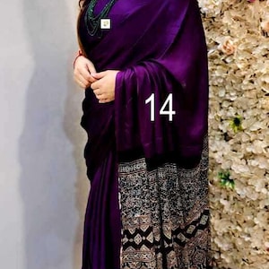 Puede incluir: Un sari de color morado intenso con un cuerpo principal de color s&oacute;lido y un borde negro en contraste con intrincados patrones geom&eacute;tricos y florales en blanco, beige y azul. La prenda incluye peque&ntilde;as borlas moradas en el dobladillo y el n&uacute;mero 14.