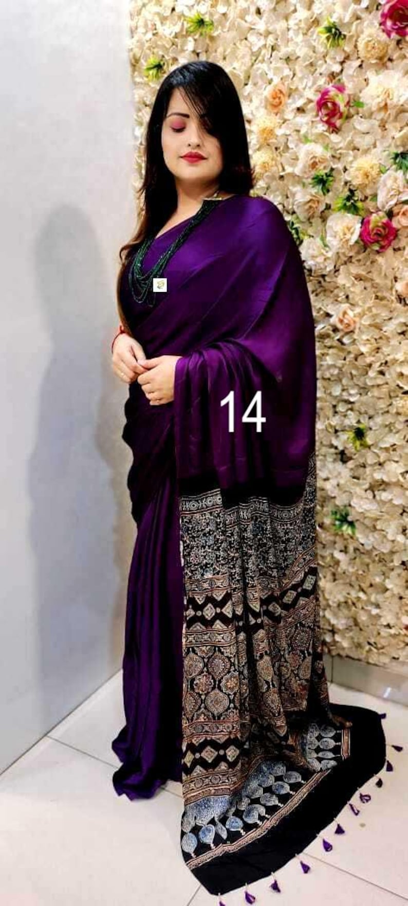Puede incluir: Un sari de color morado intenso con un cuerpo principal de color s&oacute;lido y un borde negro en contraste con intrincados patrones geom&eacute;tricos y florales en blanco, beige y azul. La prenda incluye peque&ntilde;as borlas moradas en el dobladillo y el n&uacute;mero 14.