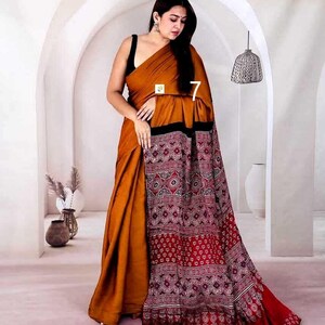 Puede incluir: Un sari de color amarillo mostaza con un pallu negro y rojo oscuro que presenta intrincados patrones de bloques estampados en blanco con motivos geom&eacute;tricos y florales. La prenda se cubre sobre una blusa negra sin mangas, mostrando una silueta larga y fluida con un borde distintivo.