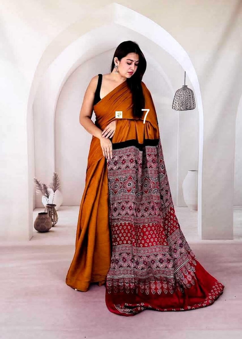 Puede incluir: Un sari de color amarillo mostaza con un pallu negro y rojo oscuro que presenta intrincados patrones de bloques estampados en blanco con motivos geom&eacute;tricos y florales. La prenda se cubre sobre una blusa negra sin mangas, mostrando una silueta larga y fluida con un borde distintivo.
