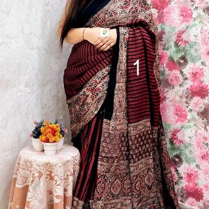 May include: A saree featuring a black bodice and a skirt with dark red and black horizontal stripes. The fabric is adorned with intricate block-printed patterns in black, red, and white, finished with a metallic gold pleated border at the hem.