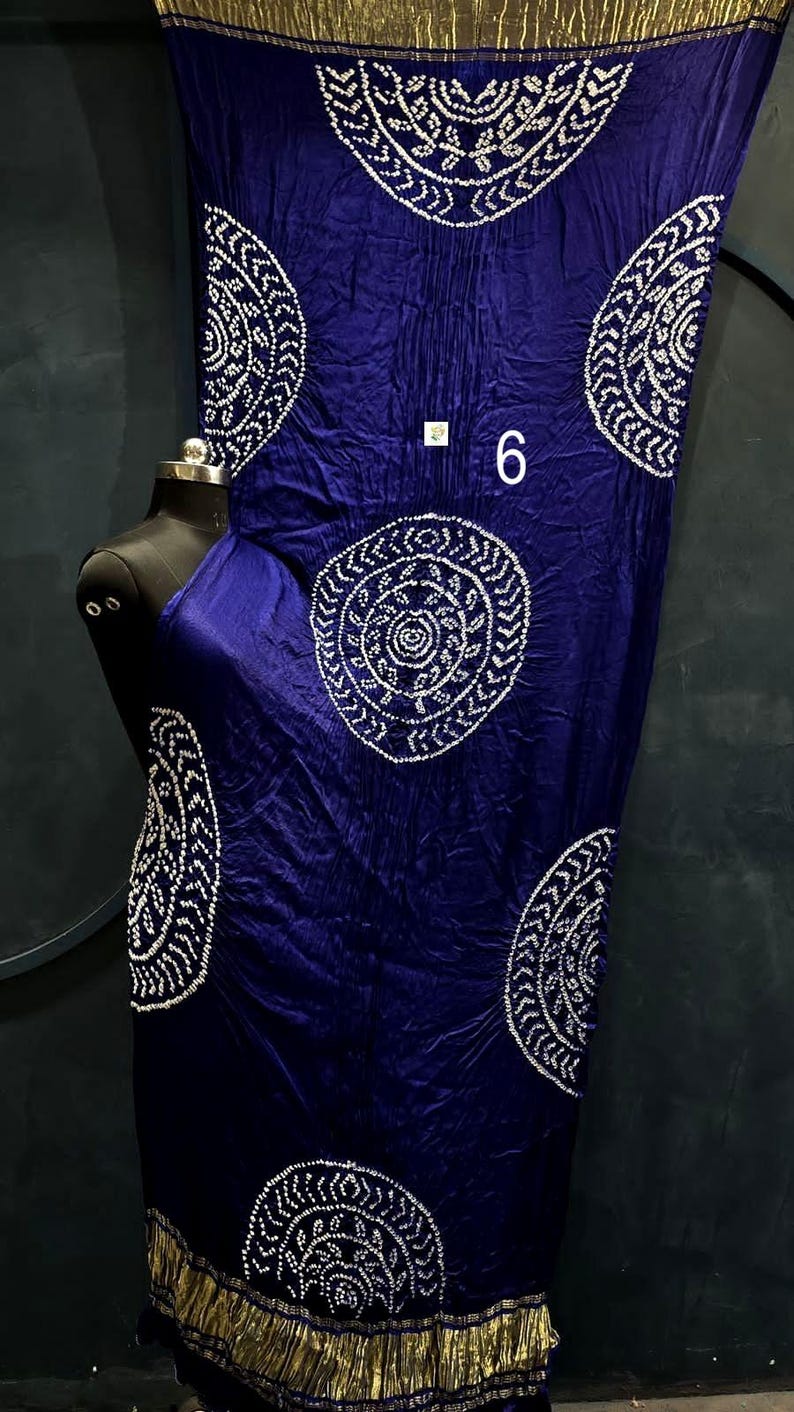 May include: A deep blue fabric drape featuring large white circular patterns with intricate dot details. The fabric has a metallic gold border at the top and bottom edges. A small white square tag with the number 6 is visible on the fabric.