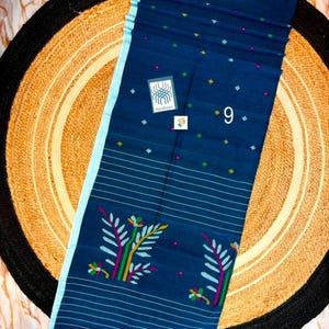 May include: A dark blue saree featuring a light blue border, thin horizontal white stripes, and small multicolored geometric motifs. The fabric displays a floral pattern in green, yellow, and pink, with a label reading Handloom and the number 9.