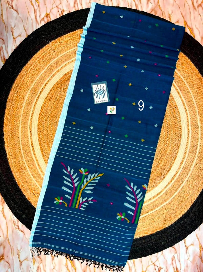 May include: A dark blue saree featuring a light blue border, thin horizontal white stripes, and small multicolored geometric motifs. The fabric displays a floral pattern in green, yellow, and pink, with a label reading Handloom and the number 9.