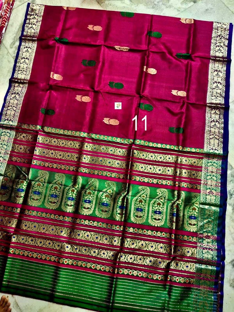 May include: A magenta saree featuring a wide green border with gold zari paisley and floral patterns. The main fabric displays small gold and green motifs scattered across the surface. The number 11 is printed in white on the center of the fabric.