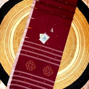 May include: A deep maroon handloom saree featuring a white striped border, small scattered white and orange dots, and two circular geometric motifs. A small white tag with the number 4 and a blue graphic logo with the text Handloom is attached.