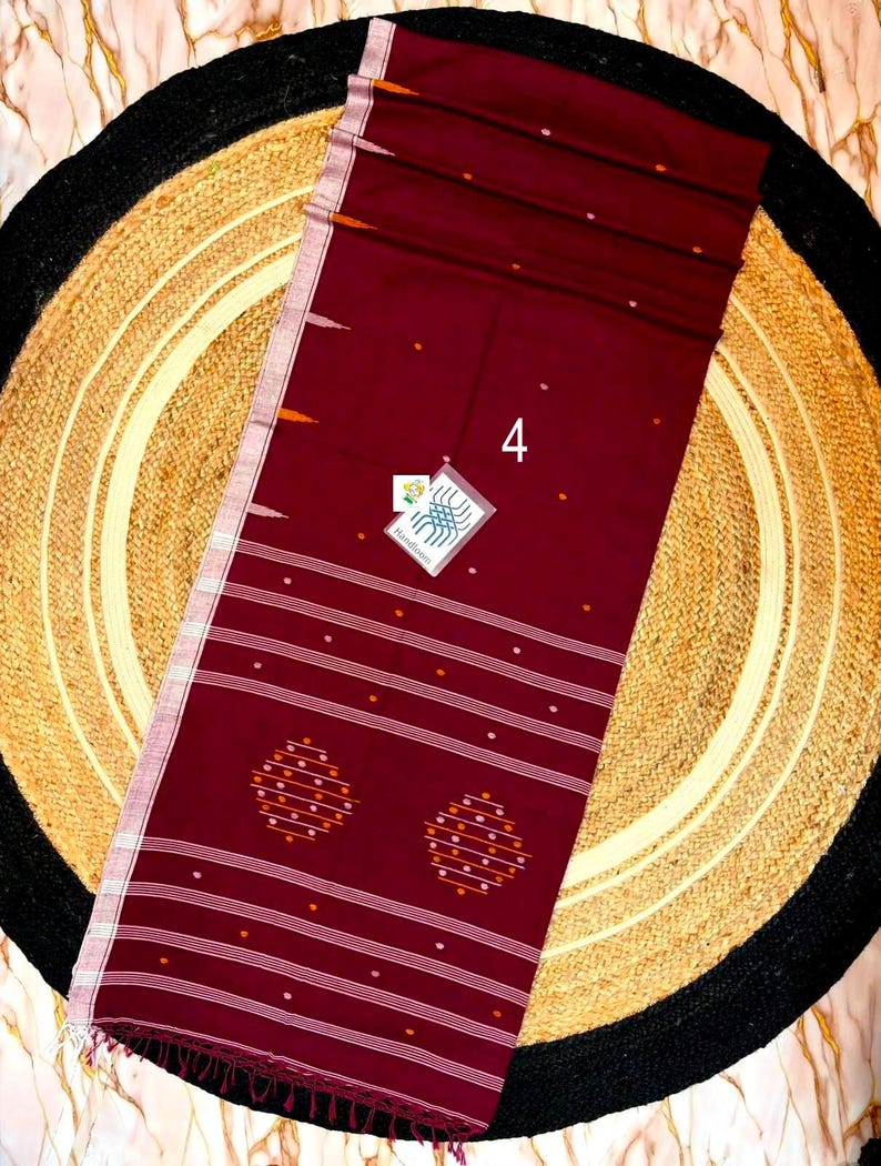 May include: A deep maroon handloom saree featuring a white striped border, small scattered white and orange dots, and two circular geometric motifs. A small white tag with the number 4 and a blue graphic logo with the text Handloom is attached.