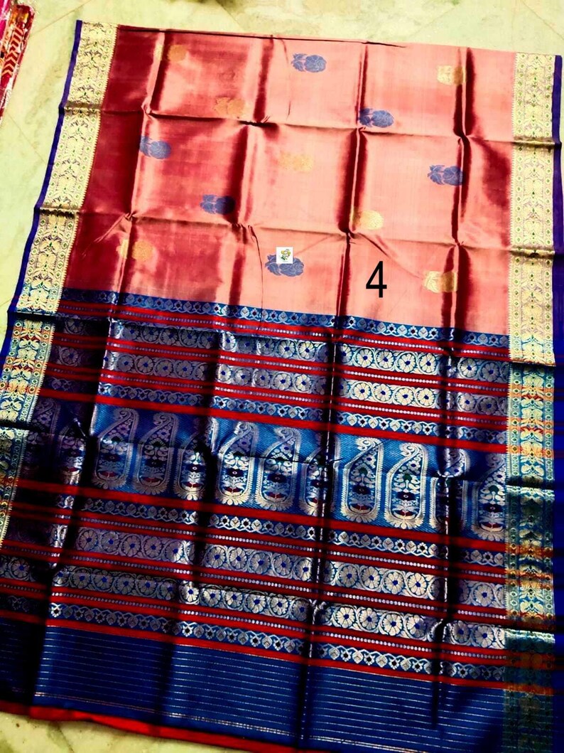 May include: A peach-colored saree featuring a large number 4 in the center, adorned with small blue and gold floral motifs. The wide border displays intricate blue and gold patterns, including paisley designs and horizontal stripes throughout.