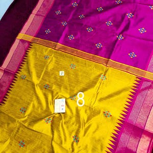 May include: A colourful textile in magenta and yellow. The fabric is decorated with small, multi-coloured floral embroidery. The design includes a yellow border with a zigzag pattern and a magenta border with stripes. Likely a saree or similar garment.
