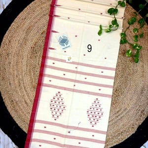 May include: A cream-colored handloom textile featuring thin horizontal red stripes and small red dot patterns. The fabric includes two larger diamond-shaped red motifs near the bottom edge, a red border, and a fringed hem. A small tag labeled 9 is visible.