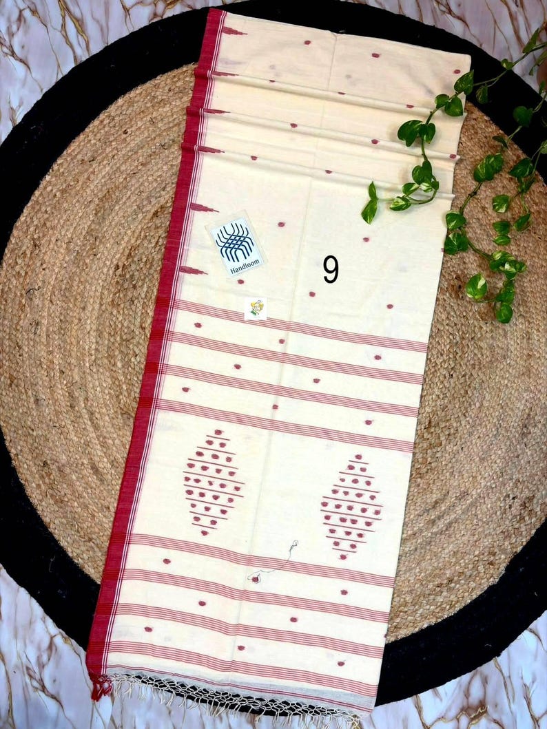 May include: A cream-colored handloom textile featuring thin horizontal red stripes and small red dot patterns. The fabric includes two larger diamond-shaped red motifs near the bottom edge, a red border, and a fringed hem. A small tag labeled 9 is visible.