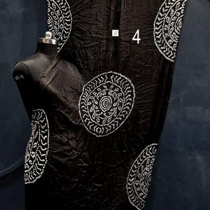 May include: A black fabric garment featuring a crinkled texture and large, circular white patterns with intricate detailing. The piece includes a metallic gold border at the top and bottom edges and displays the number 4 in white text.
