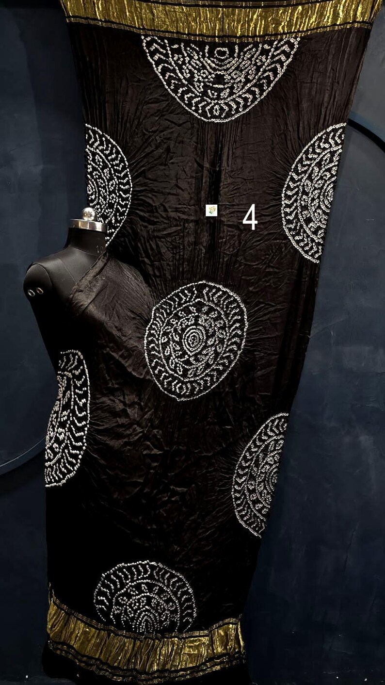 May include: A black fabric garment featuring a crinkled texture and large, circular white patterns with intricate detailing. The piece includes a metallic gold border at the top and bottom edges and displays the number 4 in white text.