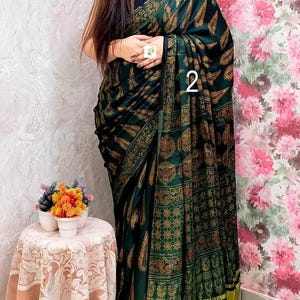 May include: A dark green saree featuring a repeating gold and brown paisley and geometric pattern, with a solid black border and a bright yellow pleated hem. The fabric is draped over the body, and the number 2 is visible on the fabric.