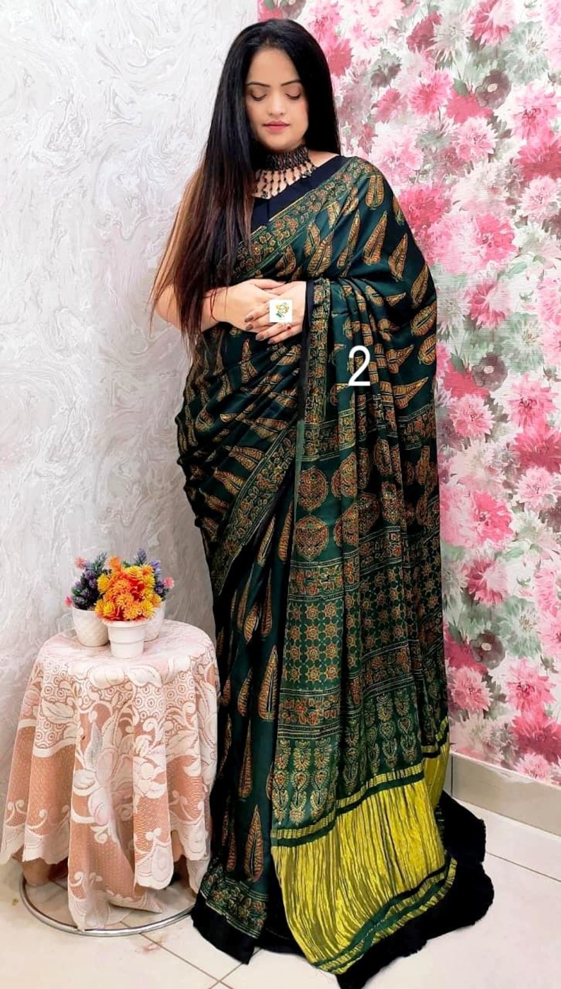 May include: A dark green saree featuring a repeating gold and brown paisley and geometric pattern, with a solid black border and a bright yellow pleated hem. The fabric is draped over the body, and the number 2 is visible on the fabric.