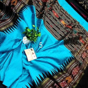 May include: A bright turquoise fabric with a wide black border featuring a repeating triangular pattern and intricate multi-colored woven motifs. The textile displays a prominent white number 5 and a small white tag attached to the center surface.