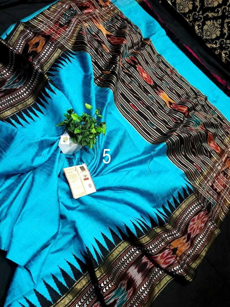 May include: A bright turquoise fabric with a wide black border featuring a repeating triangular pattern and intricate multi-colored woven motifs. The textile displays a prominent white number 5 and a small white tag attached to the center surface.