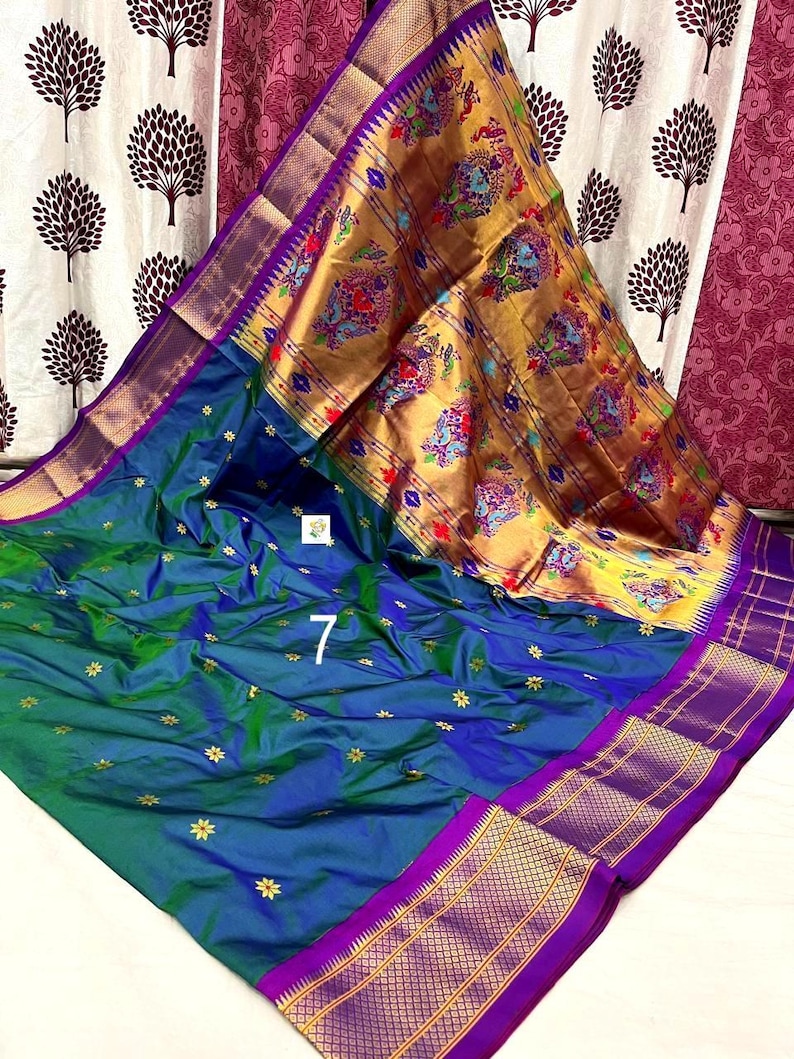 Op de afbeelding: Een blauwe en groene sari met een grote gouden pallu met kleurrijke pauwen- en bloemmotieven. Het hoofdgedeelte heeft kleine gouden stervormige patronen en de randen zijn afgewerkt met een brede paarse bies met ingewikkeld gouden zari-werk.