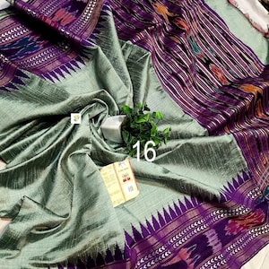 May include: A sage green saree featuring a wide purple border with intricate ikat patterns and a jagged triangular motif. The fabric has a subtle sheen and textured weave. A small white tag with the number 16 and a product label are visible.
