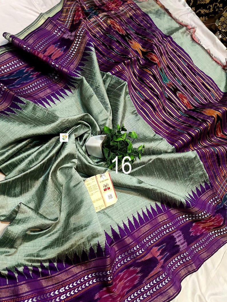 May include: A sage green saree featuring a wide purple border with intricate ikat patterns and a jagged triangular motif. The fabric has a subtle sheen and textured weave. A small white tag with the number 16 and a product label are visible.