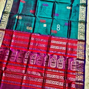 May include: A teal saree featuring small gold and purple paisley motifs, paired with a wide, vibrant red and purple border decorated with intricate gold zari patterns, floral bands, and a prominent white number 8 printed on the fabric surface.