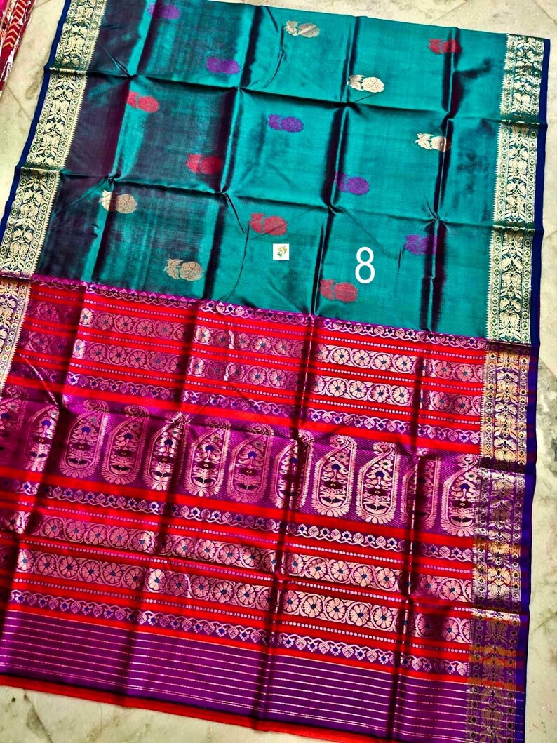 May include: A teal saree featuring small gold and purple paisley motifs, paired with a wide, vibrant red and purple border decorated with intricate gold zari patterns, floral bands, and a prominent white number 8 printed on the fabric surface.
