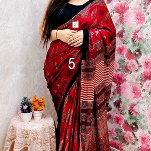 May include: A red and black saree featuring a repeating leaf pattern, a striped section, and a gold-toned fringe at the hem. The garment is paired with a black sleeveless blouse and a choker necklace. The number 5 is printed on the fabric.