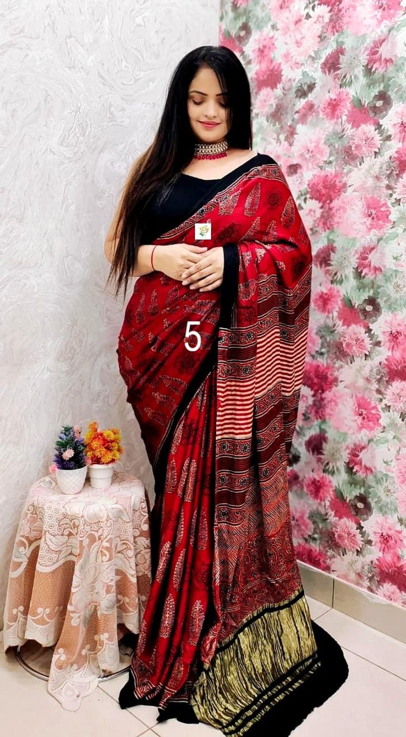 May include: A red and black saree featuring a repeating leaf pattern, a striped section, and a gold-toned fringe at the hem. The garment is paired with a black sleeveless blouse and a choker necklace. The number 5 is printed on the fabric.