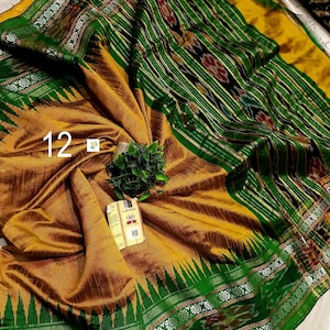 May include: A mustard yellow and emerald green saree featuring intricate ikat patterns, a wide border with geometric motifs, and a decorative pallu. The fabric displays a textured weave with the number 12 and a small product tag attached.
