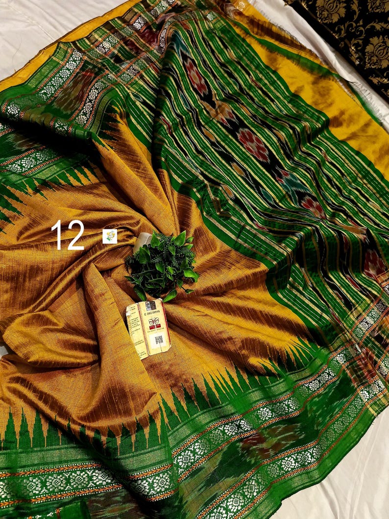 May include: A mustard yellow and emerald green saree featuring intricate ikat patterns, a wide border with geometric motifs, and a decorative pallu. The fabric displays a textured weave with the number 12 and a small product tag attached.