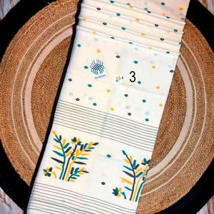 May include: A cream-colored handloom saree featuring a scattered pattern of small teal and yellow dots. The fabric includes a black border, horizontal black stripes, and a large botanical print in yellow and teal. A label reads Handloom and 3.