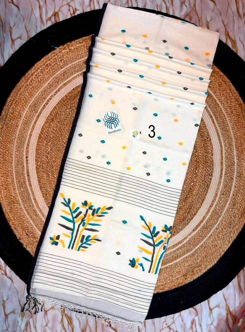 May include: A cream-colored handloom saree featuring a scattered pattern of small teal and yellow dots. The fabric includes a black border, horizontal black stripes, and a large botanical print in yellow and teal. A label reads Handloom and 3.