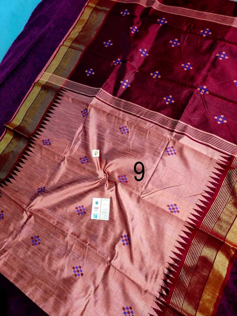 May include: A maroon and peach-coloured silk sari with a geometric pattern of small blue and purple squares. The sari has a decorative border with gold accents and a zigzag edge. The number "9" is visible on the sari.