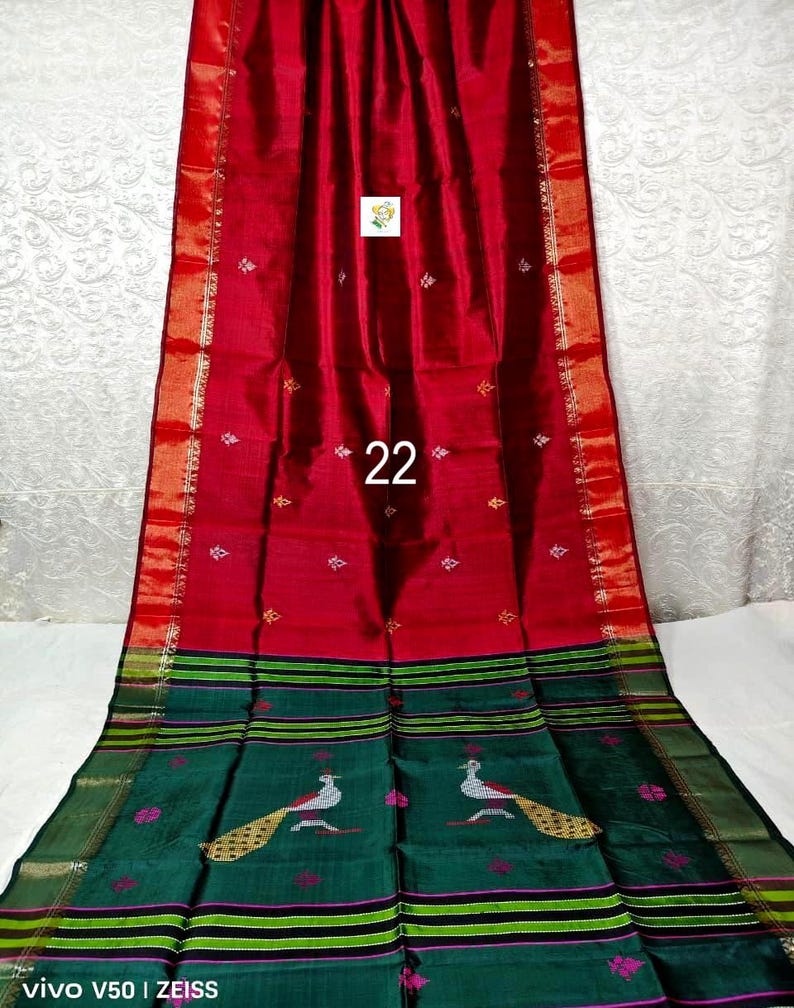 Puede incluir: Un sari de color rojo intenso con un borde ancho de color rojo vibrante y detalles met&aacute;licos dorados. La secci&oacute;n inferior es un panel verde oscuro decorado con motivos de pavos reales en blanco y oro, con rayas horizontales negras y verdes.