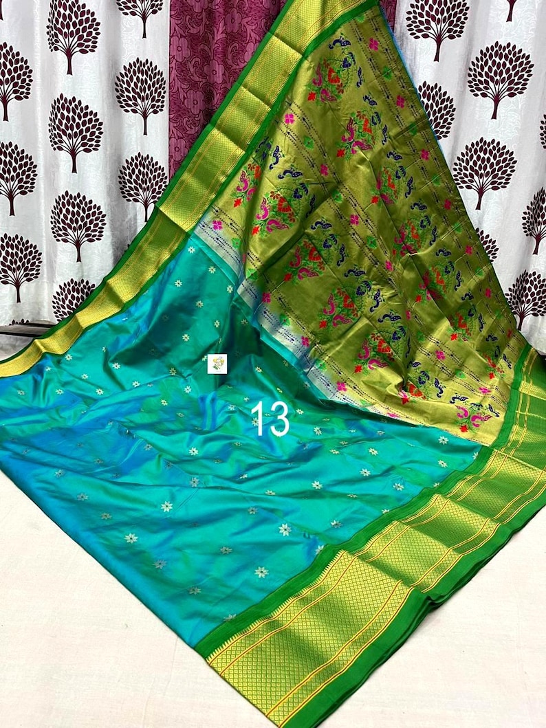 Op de afbeelding: Een turquoise sari met kleine zilveren bloemmotieven over het hele lichaam, gecombineerd met een brede olijfgroene rand en een pallu versierd met ingewikkelde pauwen- en bloempatronen in roze, groen en blauw, gemarkeerd met het nummer 13.